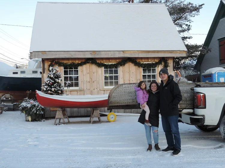 The restored red Ackroyd skiff loaded on a truck in the snow with a family standing beside it for delivery to the Seripham HomeSchool