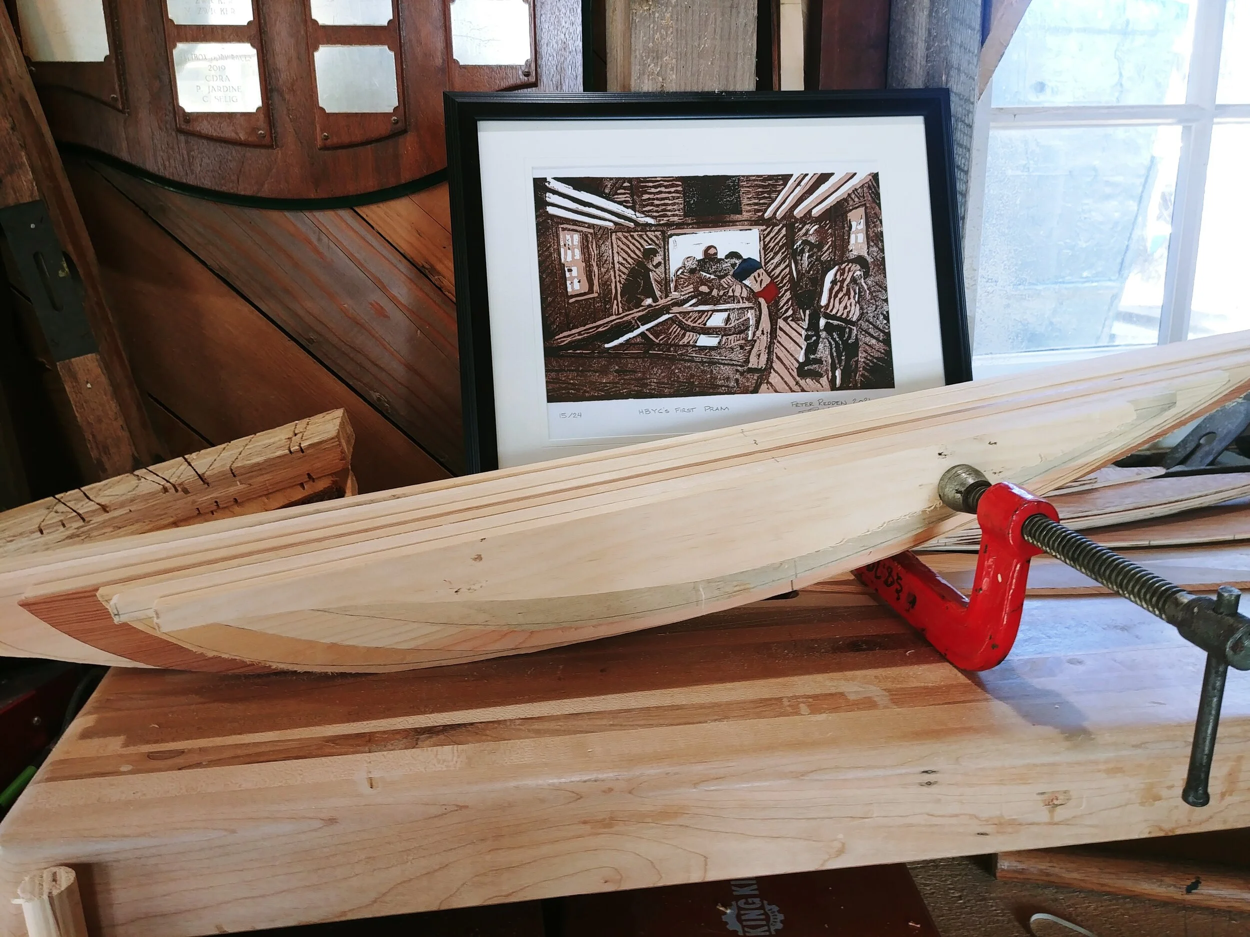 A half hull model blank glued up and clamped on the workbench beside a framed print of the Co-op's first pram