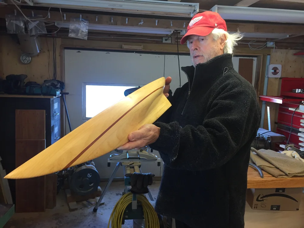 A workshop participant holding a finished light-wood half hull with a contrasting dark keel stripe in the boat shed