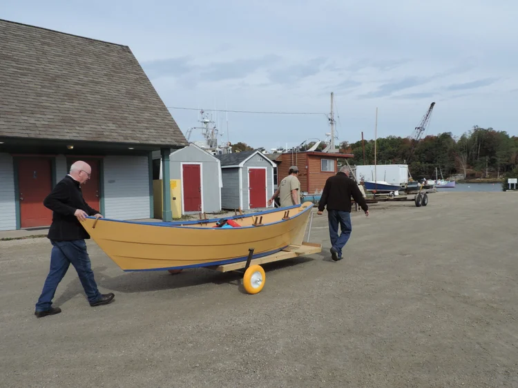 Two volunteers wheeling the freshly restored yellow LaHave Dory across the Boatyard between sheds