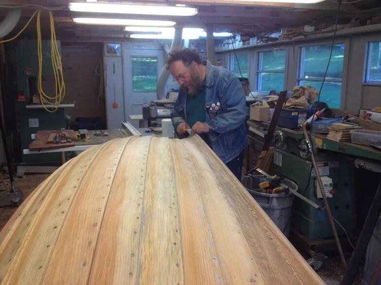 A boatbuilder working on the bare-wood planking of the upturned Lawley tender hull in the Co-op shop