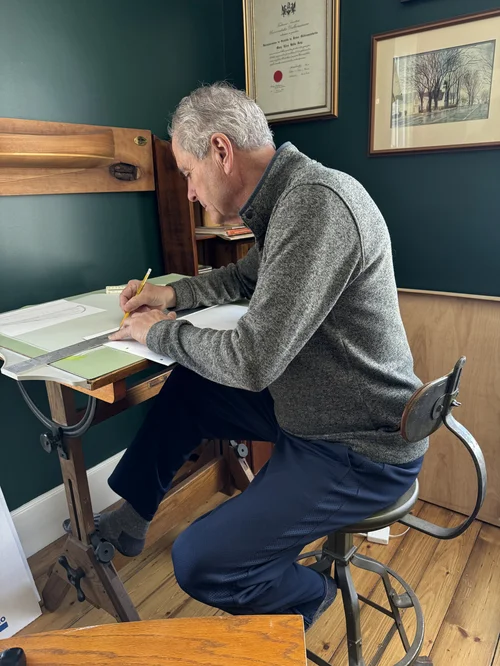 Peter at William Roue's antique drafting table and stool, drawing the Mush-a-Mush skiff construction plans in his home office