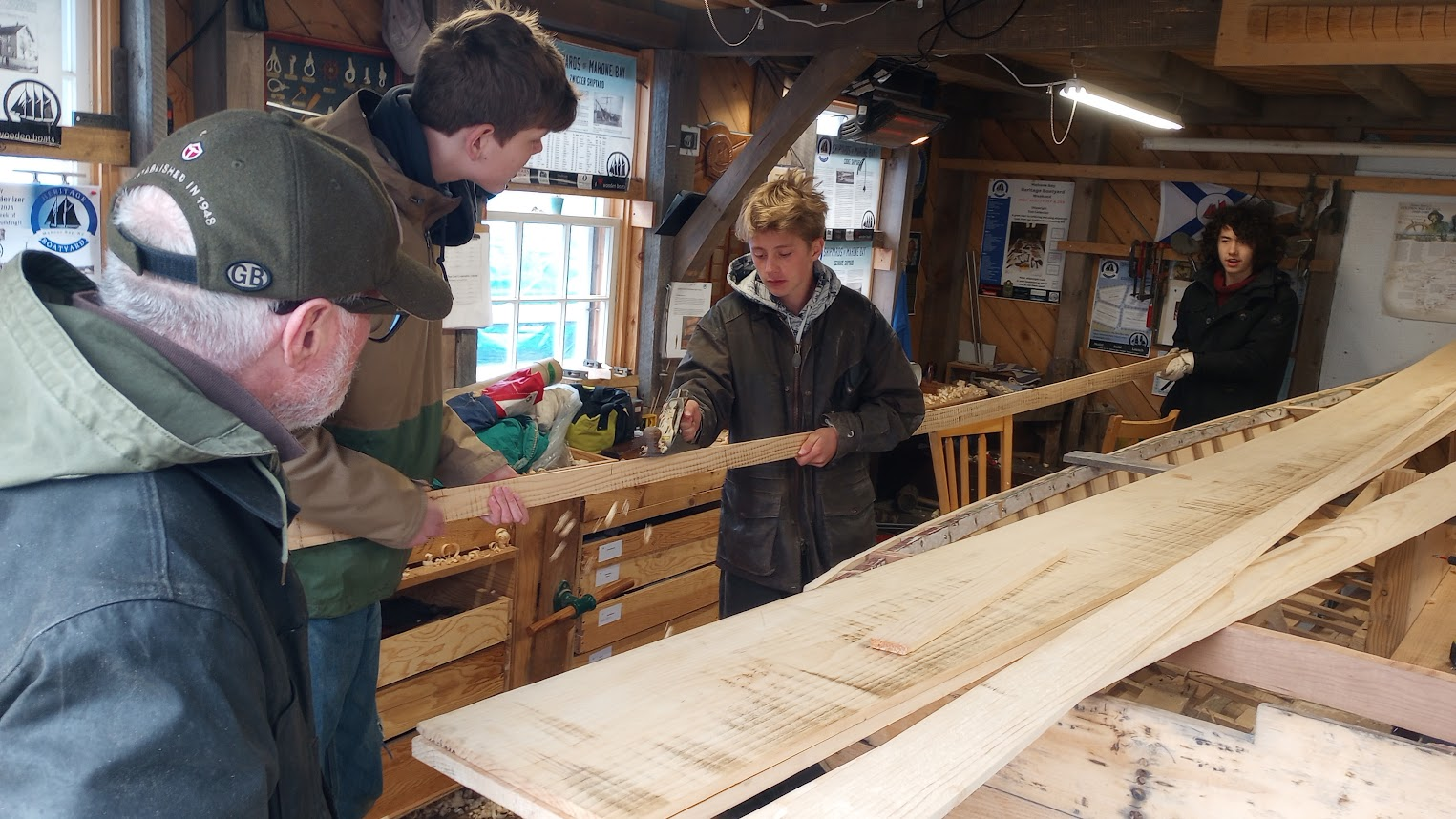 Volunteers and teenage students working on a long Pintail Skiff plank inside the Heritage Boatyard shed