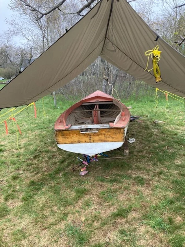 The Pintail Skiff bow-on under a tan tarp shelter, faded yellow and red paint still on the hull before stripping