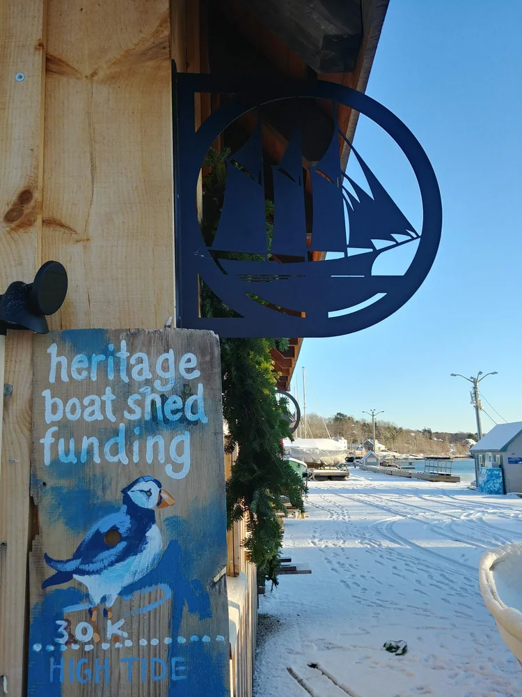 Painted 'Heritage Boat Shed Funding — 30K High Tide' puffin sign mounted on the shed beneath the schooner silhouette emblem