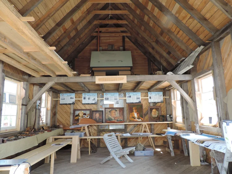 Inside the shed — exposed timber rafters with framed shipwright portraits and Shipyards of Mahone Bay panels along the wall