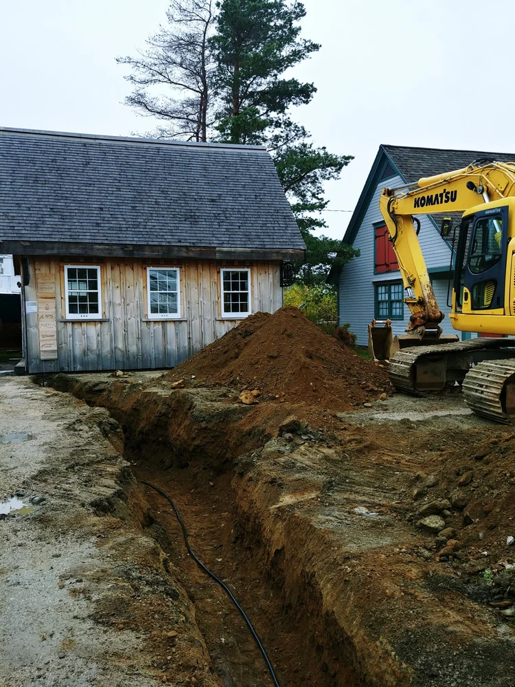 An excavator digging a utilities trench from the boatbuilding shed across the property