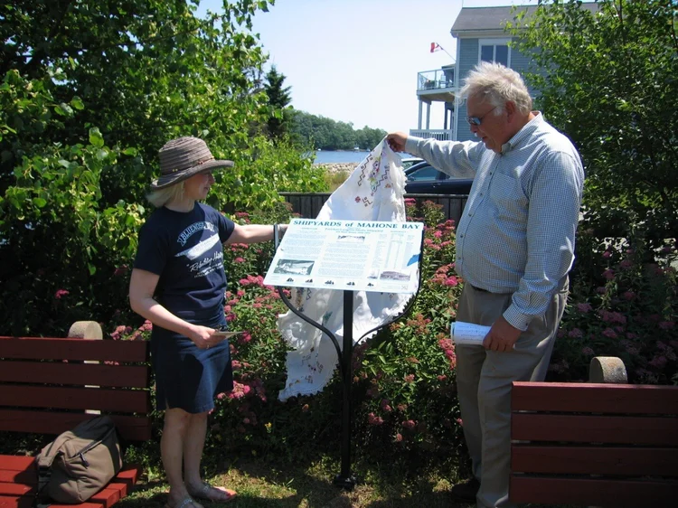 Two volunteers unveiling a Shipyards of Mahone Bay interpretive sign on its post by the waterfront