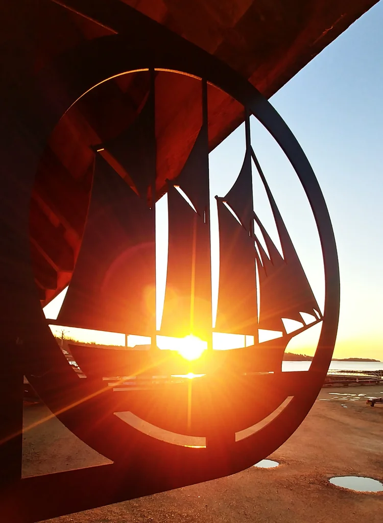 A black metal sailing ship silhouette sign backlit by a low sun on the Mahone Bay waterfront