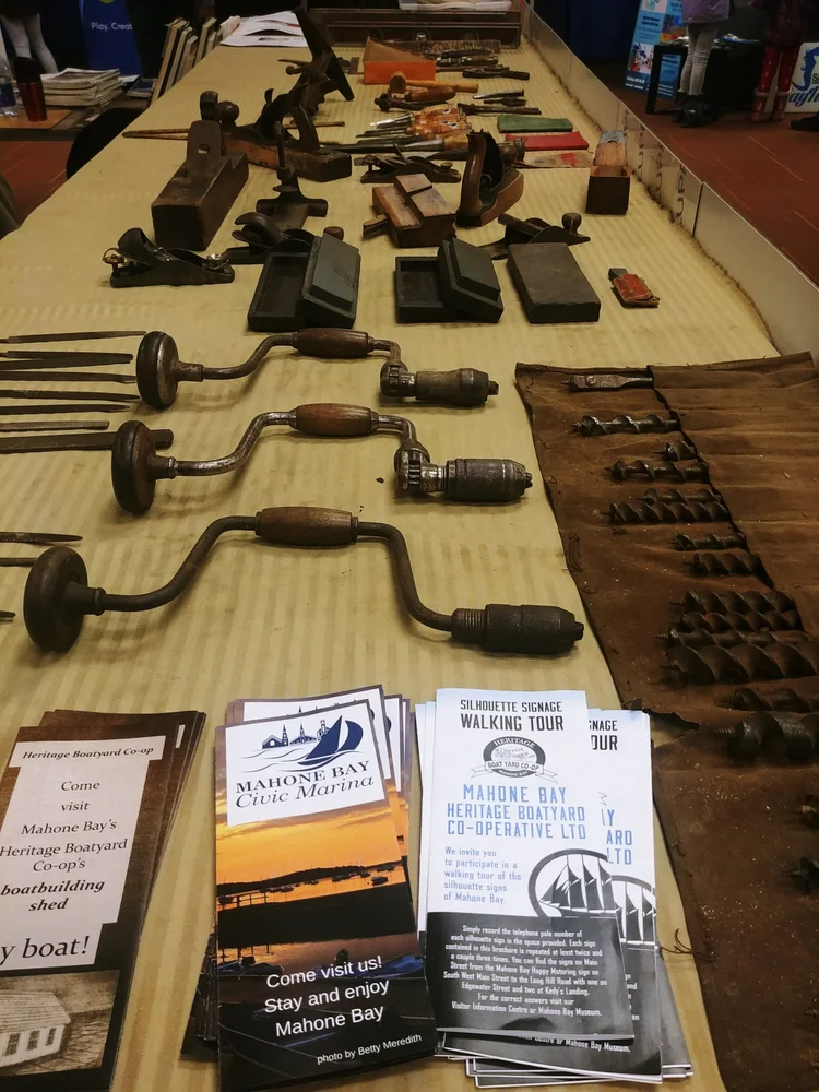 Long table displaying donated antique boatbuilding tools — hand drills, planes, augers, and brochures