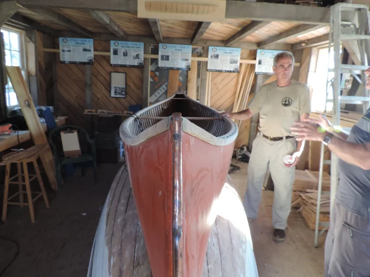 Bow-on view of the Tremblay canoe partway through restoration in the Co-op shed with a volunteer alongside